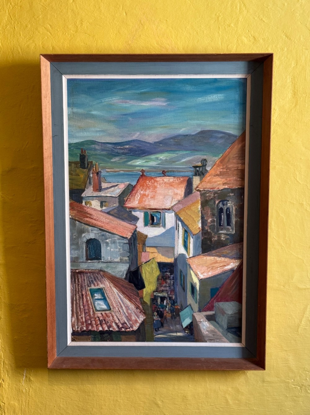 A mid-century oil painting on board depicting a narrow street in Antibes with terracotta roofs and distant mountains, housed in a grey and timber wooden frame.