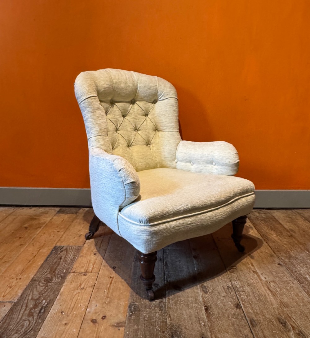A Victorian armchair upholstered in cream fabric featuring deep button tufting on the backrest, padded arms, and dark turned wooden legs with brass castors.