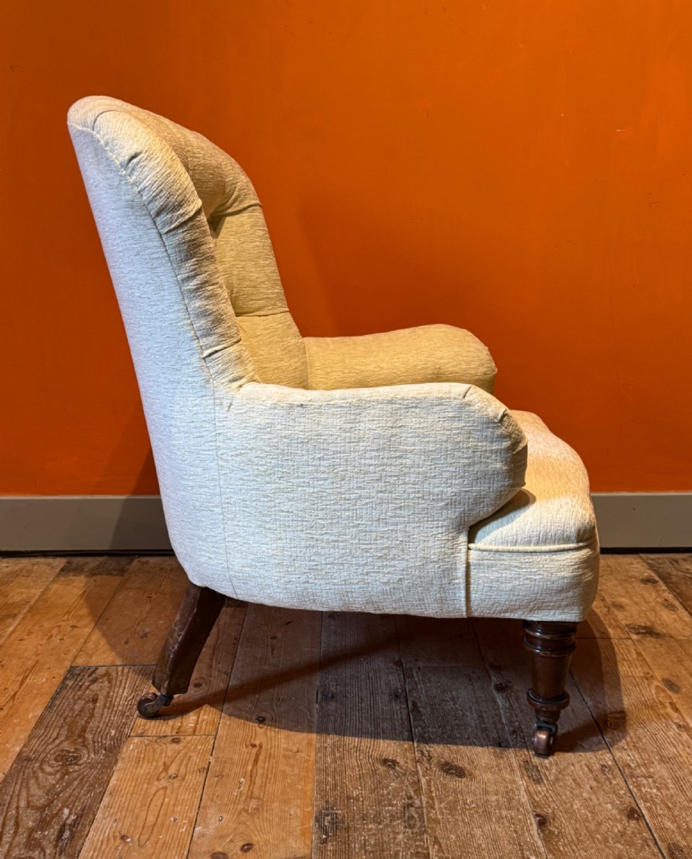 A Victorian armchair upholstered in cream fabric featuring deep button tufting on the backrest, padded arms, and dark turned wooden legs with brass castors. View V