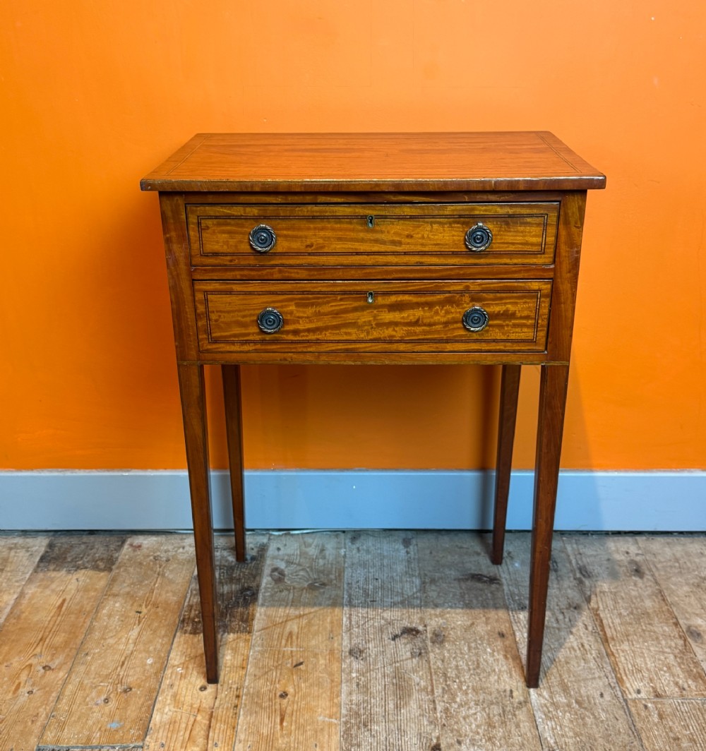 A Sheraton style satinwood side table featuring two crossbanded drawers with round brass pulls and square tapered legs.
