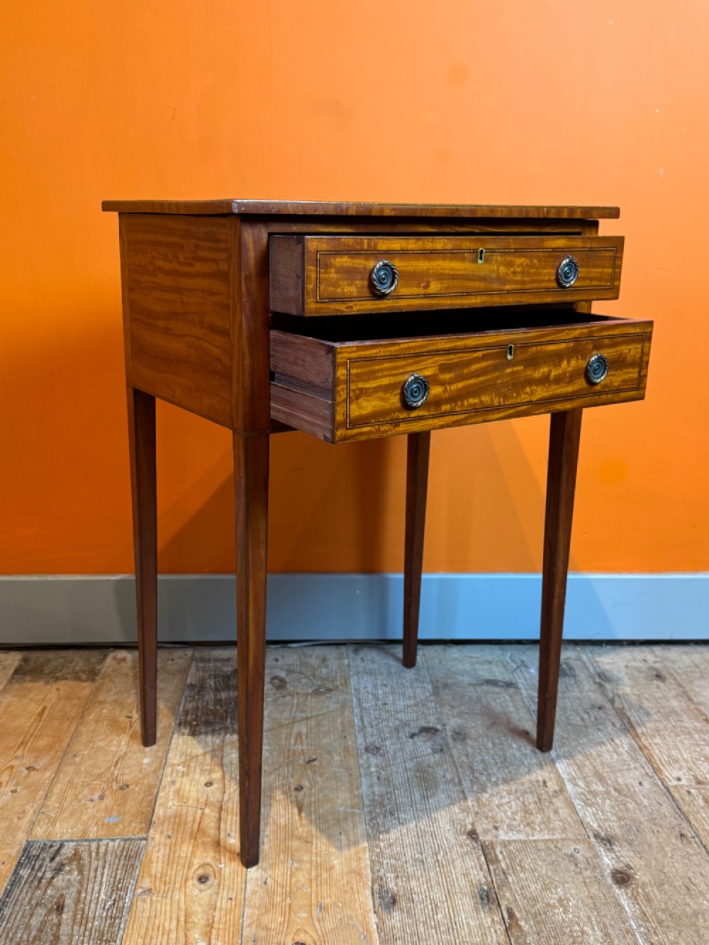 A Sheraton style satinwood side table featuring two crossbanded drawers with round brass pulls and square tapered legs. View IV