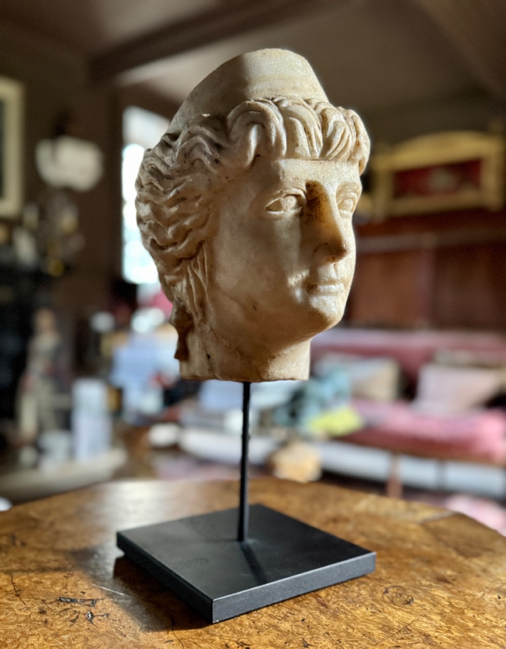 A carved white marble head of a classical goddess wearing a diadem, mounted on a slender dark metal display stand. View II