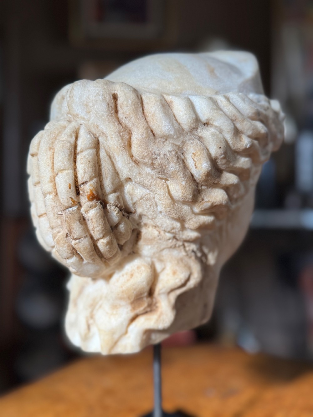 A carved white marble head of a classical goddess wearing a diadem, mounted on a slender dark metal display stand. View IV