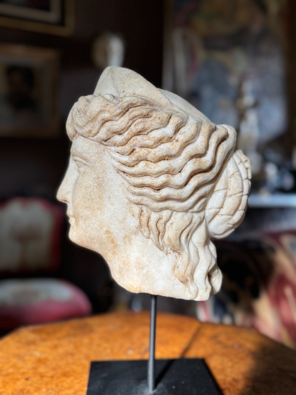 A carved white marble head of a classical goddess wearing a diadem, mounted on a slender dark metal display stand. View VI