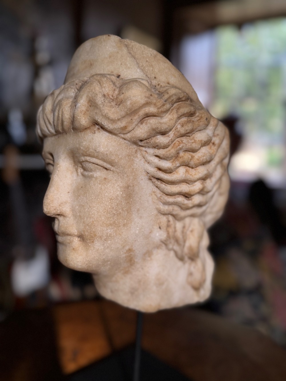 A carved white marble head of a classical goddess wearing a diadem, mounted on a slender dark metal display stand. View VII