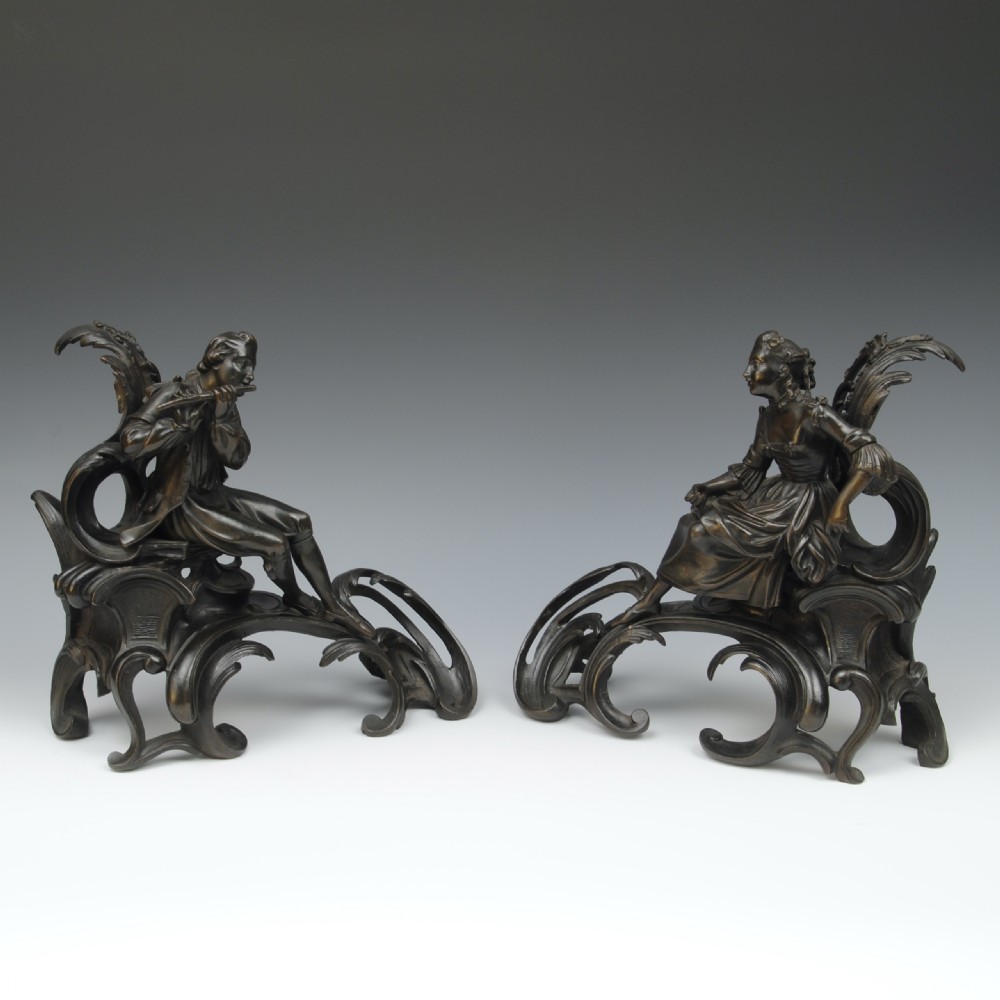 A pair of dark patinated bronze 19th-century French Rococo style chenets featuring sculpted figures seated atop elaborate scrollwork bases.