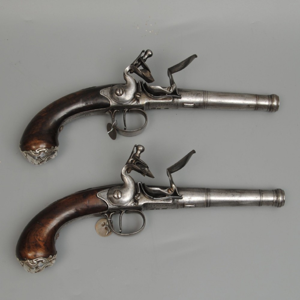A matched pair of antique Queen Anne flintlock pistols featuring wooden stocks, steel barrels, and intricate silver mounts against a neutral background.