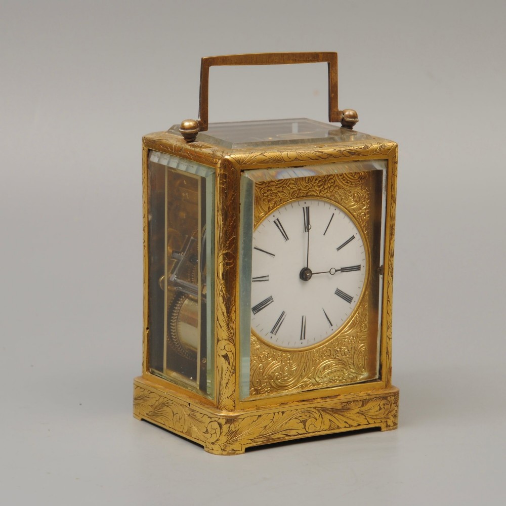 A small rectangular gilt brass carriage clock decorated with foliate engraving, bevelled glass panels, a top handle, and a white enamel dial with Roman numerals.