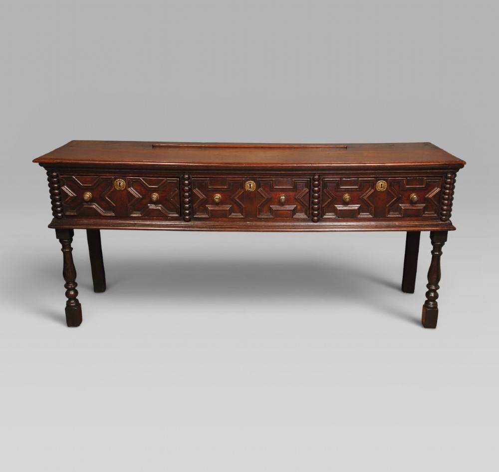A late 17th-century dark oak dresser base featuring three drawers with geometric mouldings, brass knobs, and turned front legs.