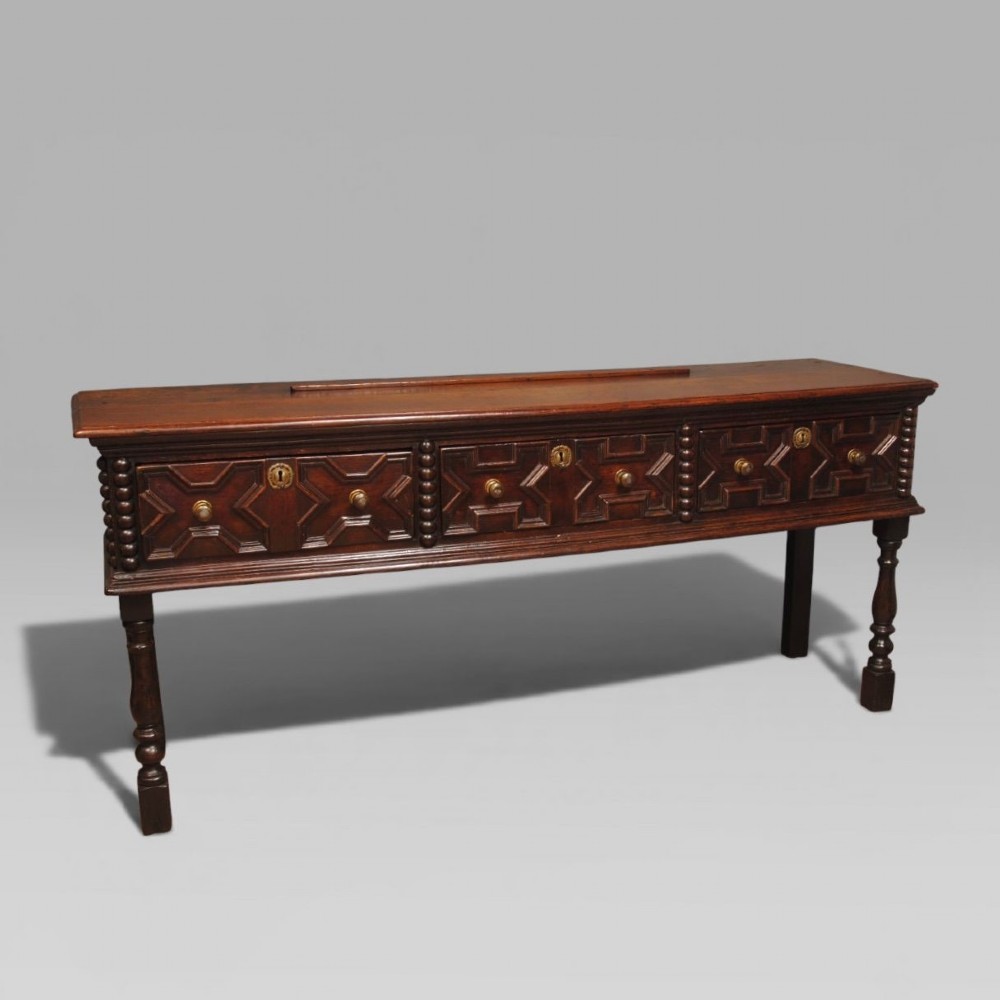 A late 17th-century dark oak dresser base featuring three drawers with geometric mouldings, brass knobs, and turned front legs. View II