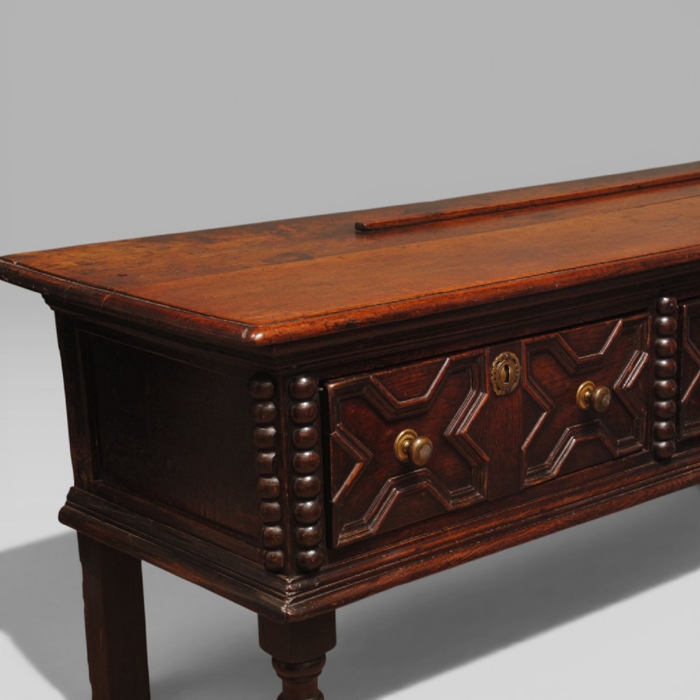 A late 17th-century dark oak dresser base featuring three drawers with geometric mouldings, brass knobs, and turned front legs. View III
