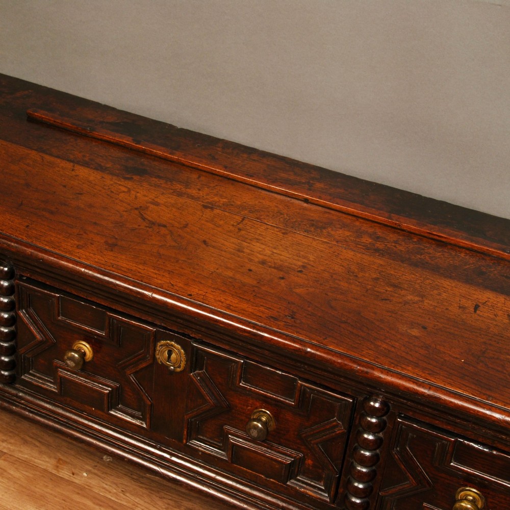 A late 17th-century dark oak dresser base featuring three drawers with geometric mouldings, brass knobs, and turned front legs. View IV