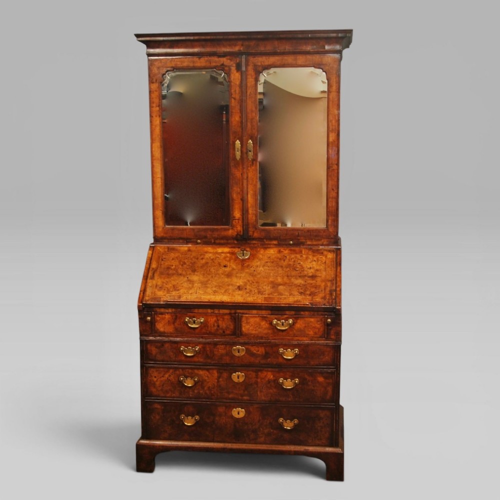 Queen Anne burr walnut bureau bookcase featuring upper double arched mirrored doors above a slant-front writing desk and graduated lower drawers with brass handles.