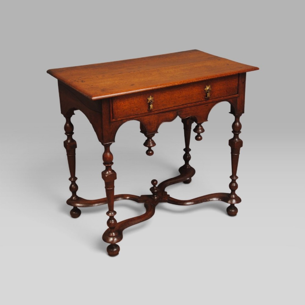 An eighteenth-century Queen Anne oak lowboy featuring turned legs, a single drawer with brass pulls, a shaped apron with drop finials, and a curved X-stretcher.
