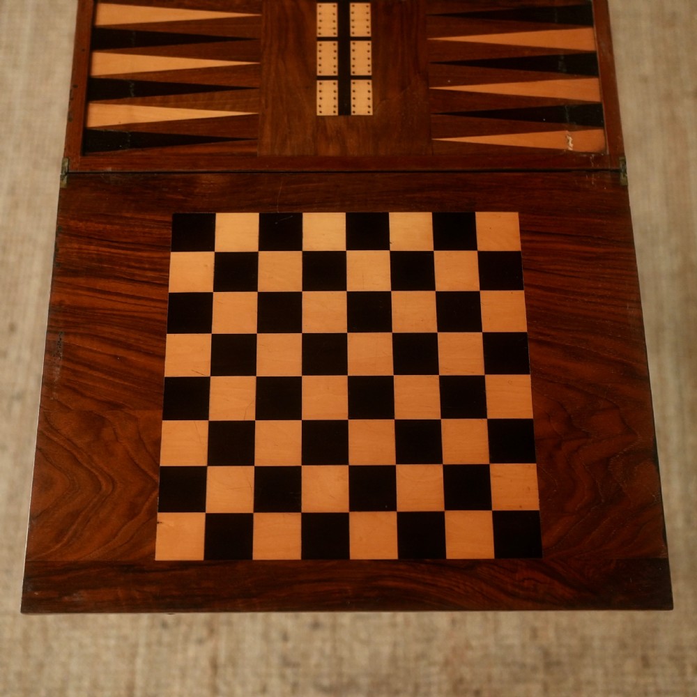 A nineteenth-century rosewood games table featuring a swivel folding top with inlaid chess and backgammon boards, resting on turned trestle supports with a fabric sewing basket underneath. View II