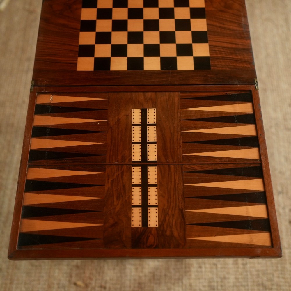 A nineteenth-century rosewood games table featuring a swivel folding top with inlaid chess and backgammon boards, resting on turned trestle supports with a fabric sewing basket underneath. View IV