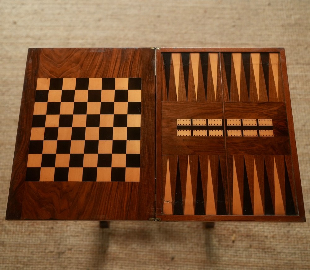 A nineteenth-century rosewood games table featuring a swivel folding top with inlaid chess and backgammon boards, resting on turned trestle supports with a fabric sewing basket underneath. View V