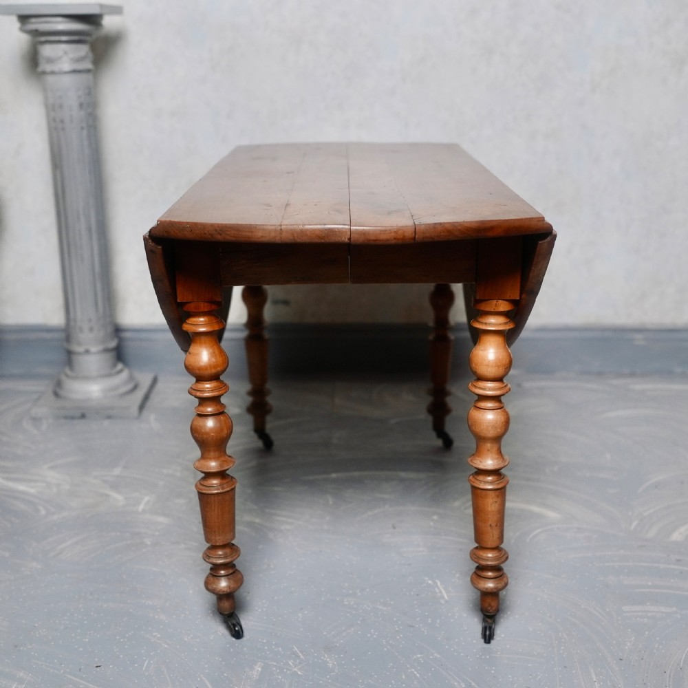An antique French fruitwood drop-leaf dining table with turned legs, castors, and a warm honey-toned wood grain. View II