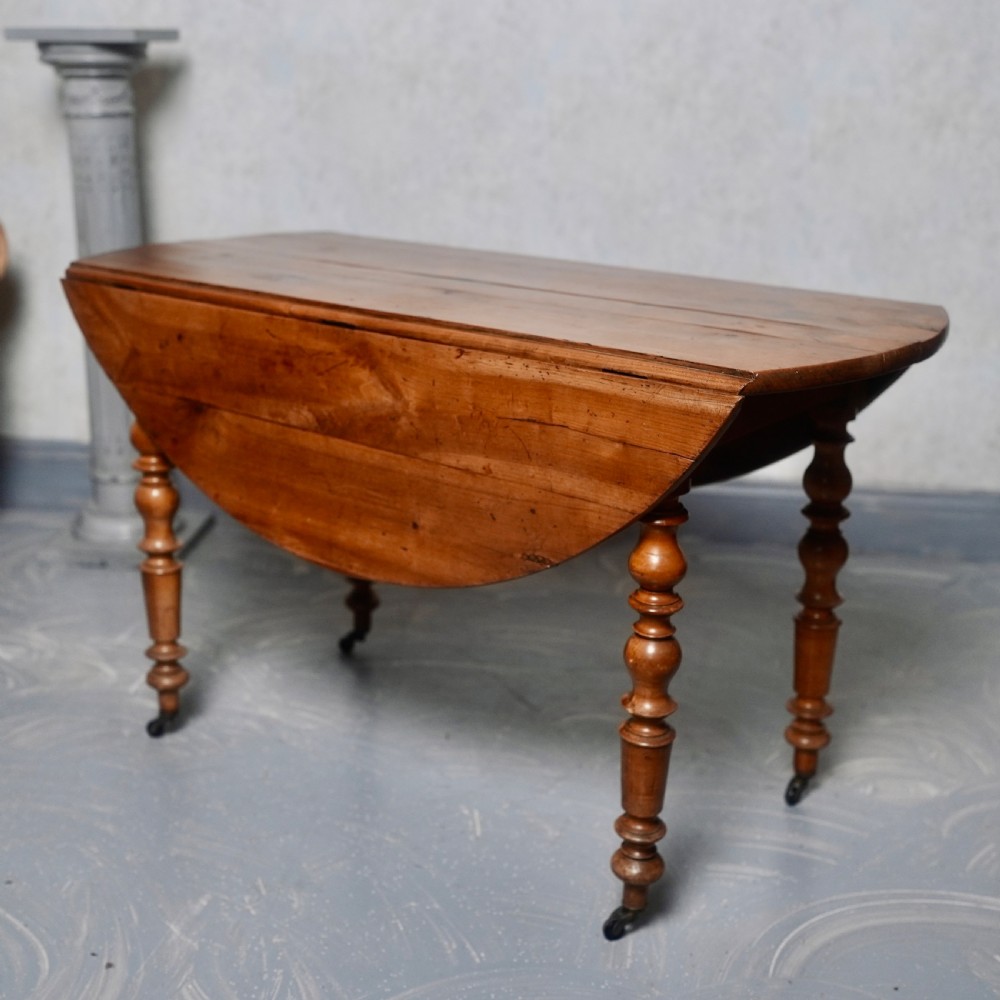 An antique French fruitwood drop-leaf dining table with turned legs, castors, and a warm honey-toned wood grain. View III