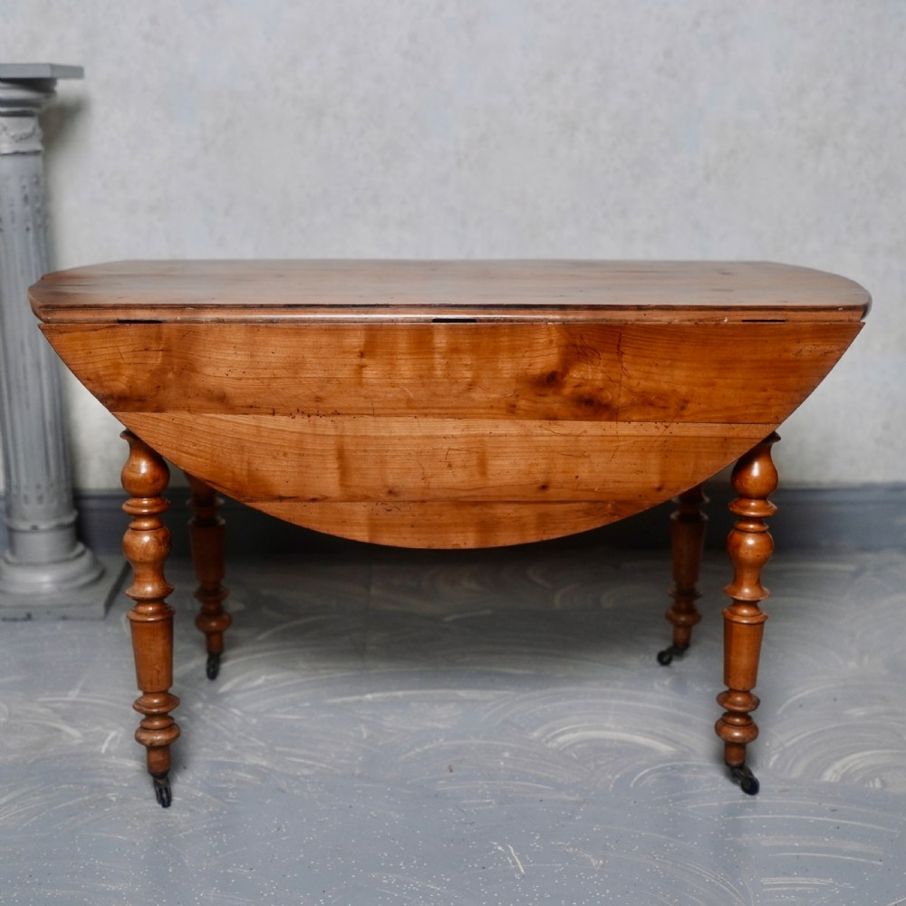 An antique French fruitwood drop-leaf dining table with turned legs, castors, and a warm honey-toned wood grain. View IV