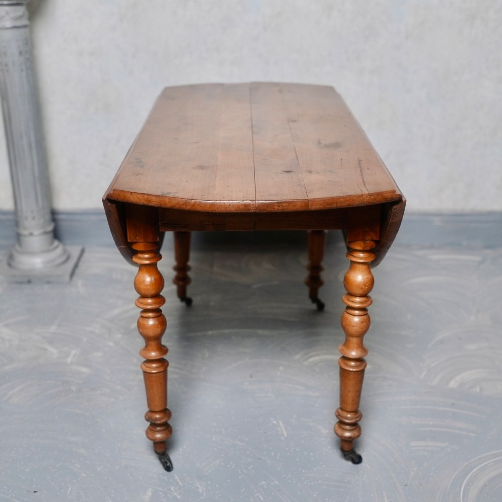 An antique French fruitwood drop-leaf dining table with turned legs, castors, and a warm honey-toned wood grain. View V