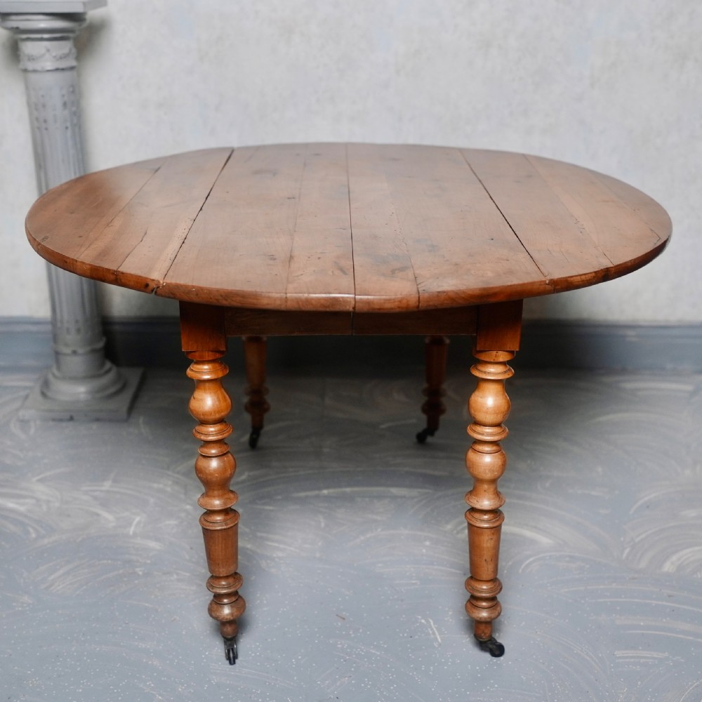 An antique French fruitwood drop-leaf dining table with turned legs, castors, and a warm honey-toned wood grain. View VII