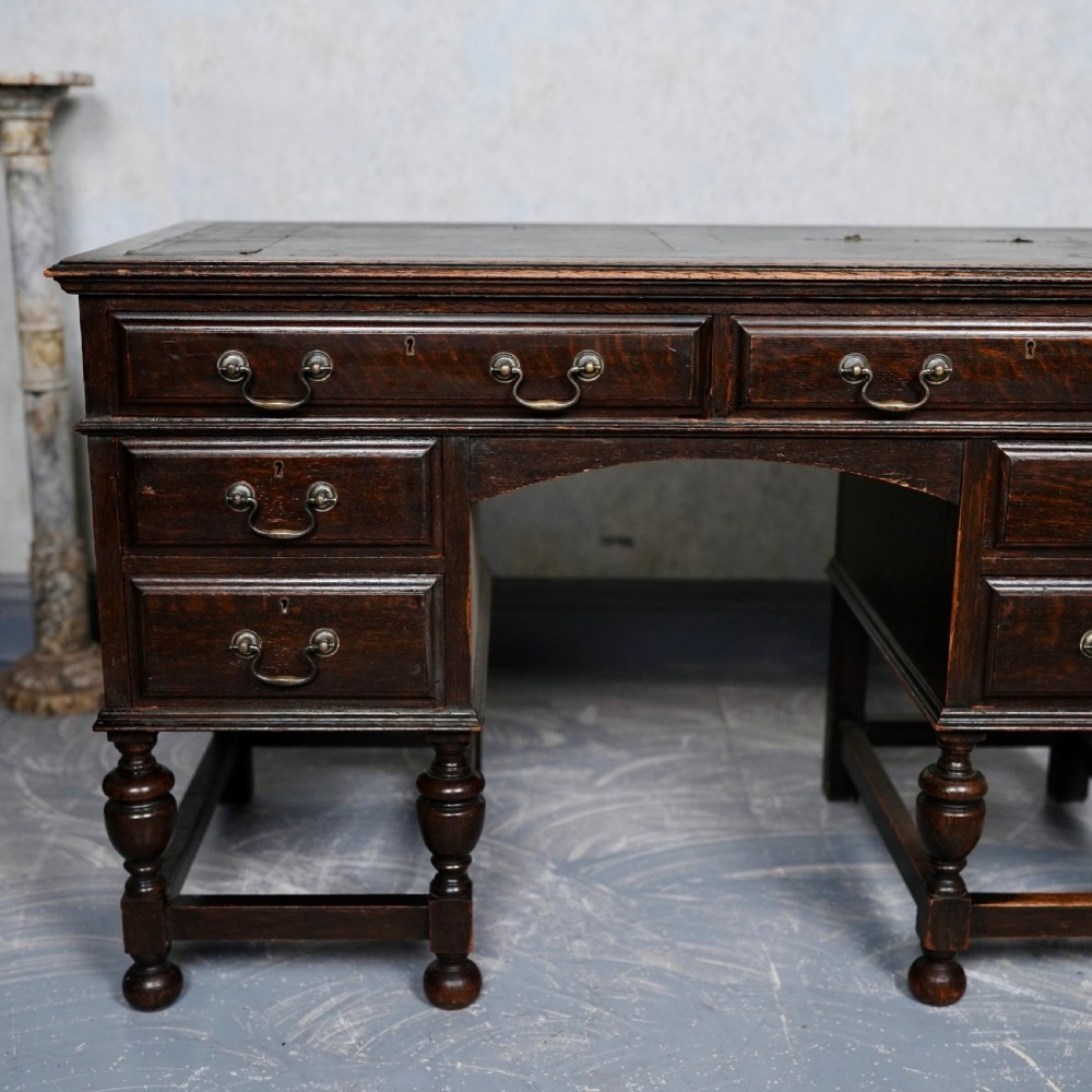 An antique dark oak kneehole desk featuring a leather-inset writing surface, six drawers with brass handles, and turned legs connected by stretchers. View II