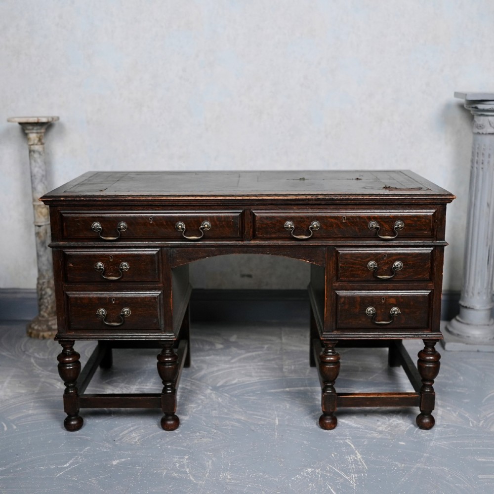 An antique dark oak kneehole desk featuring a leather-inset writing surface, six drawers with brass handles, and turned legs connected by stretchers.