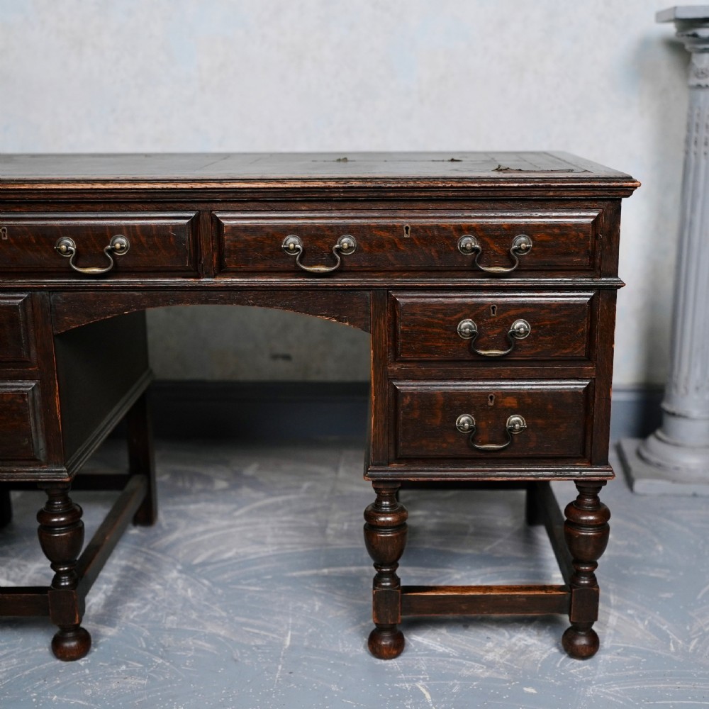 An antique dark oak kneehole desk featuring a leather-inset writing surface, six drawers with brass handles, and turned legs connected by stretchers. View IV
