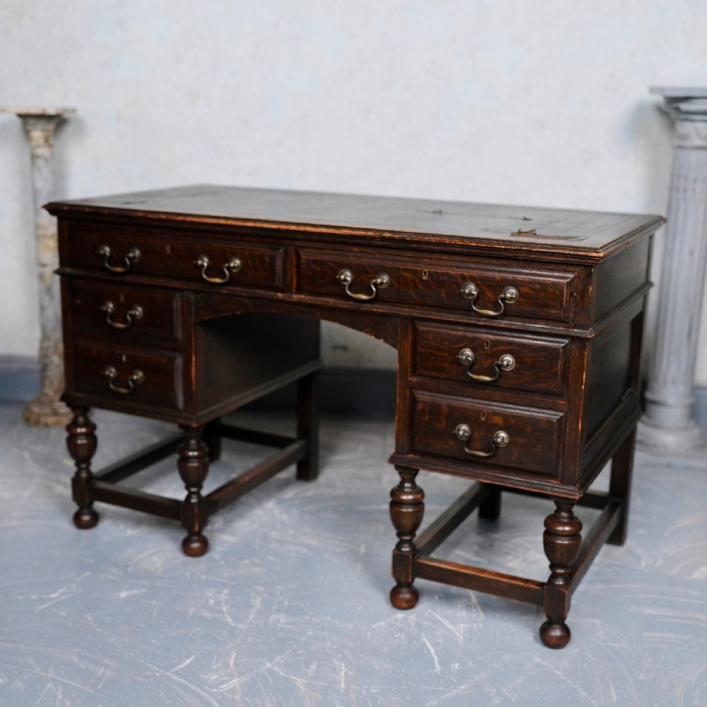 An antique dark oak kneehole desk featuring a leather-inset writing surface, six drawers with brass handles, and turned legs connected by stretchers. View VII