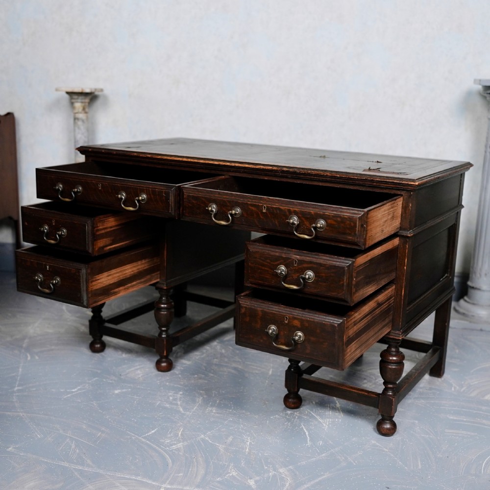 An antique dark oak kneehole desk featuring a leather-inset writing surface, six drawers with brass handles, and turned legs connected by stretchers. View VIII
