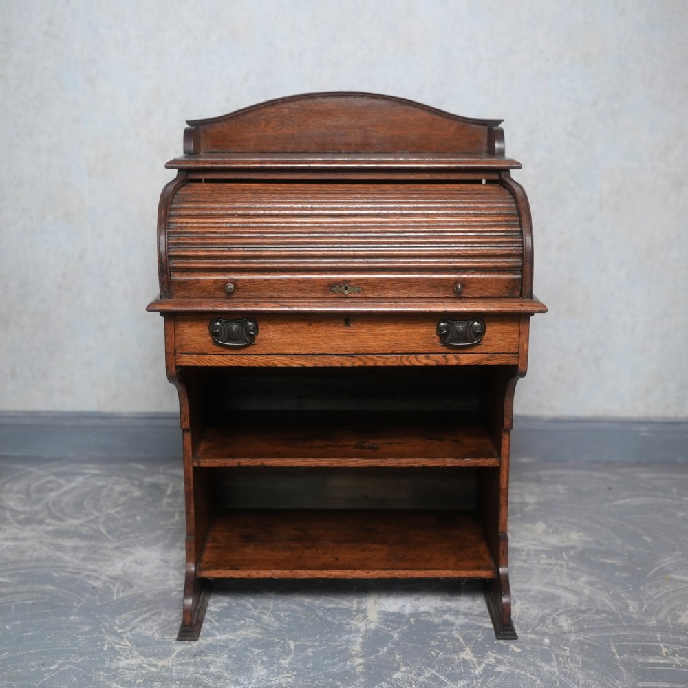An Edwardian oak rolltop writing bureau featuring a tambour slatted shutter, a single frieze drawer with dark metal handles, an arched top gallery, and two open lower shelves.