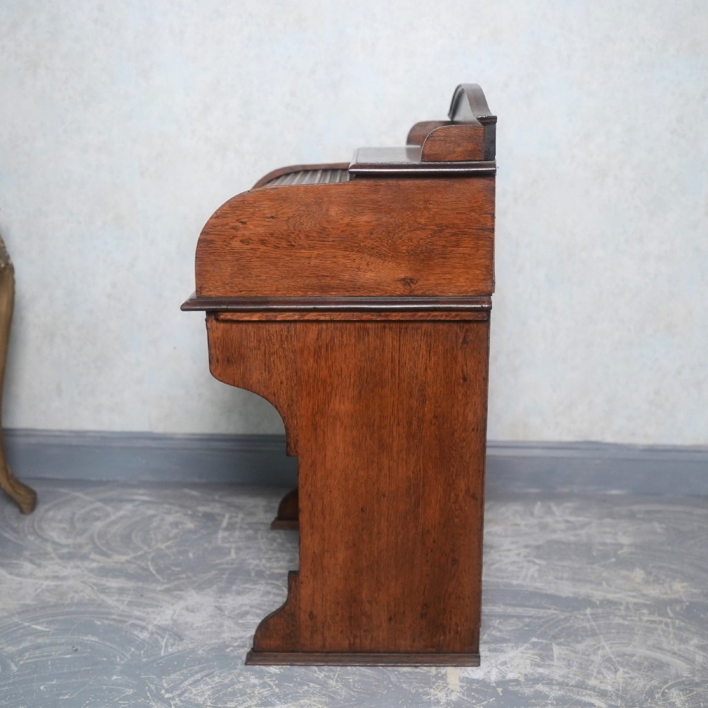 An Edwardian oak rolltop writing bureau featuring a tambour slatted shutter, a single frieze drawer with dark metal handles, an arched top gallery, and two open lower shelves. View IX