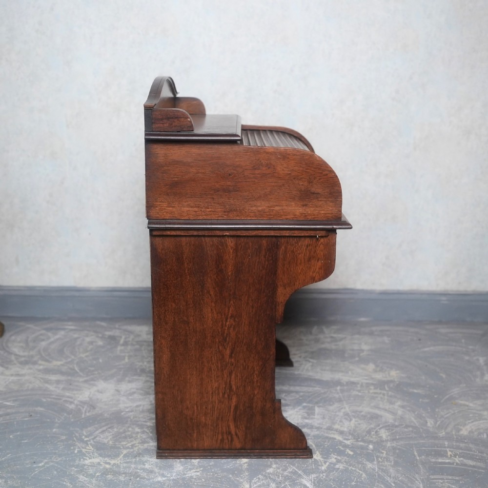 An Edwardian oak rolltop writing bureau featuring a tambour slatted shutter, a single frieze drawer with dark metal handles, an arched top gallery, and two open lower shelves. View XI