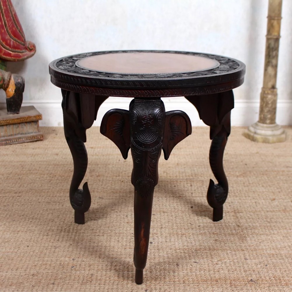 A circular dark hardwood accent table featuring a carved relief border and three legs shaped like elephant heads with downward curving trunks.