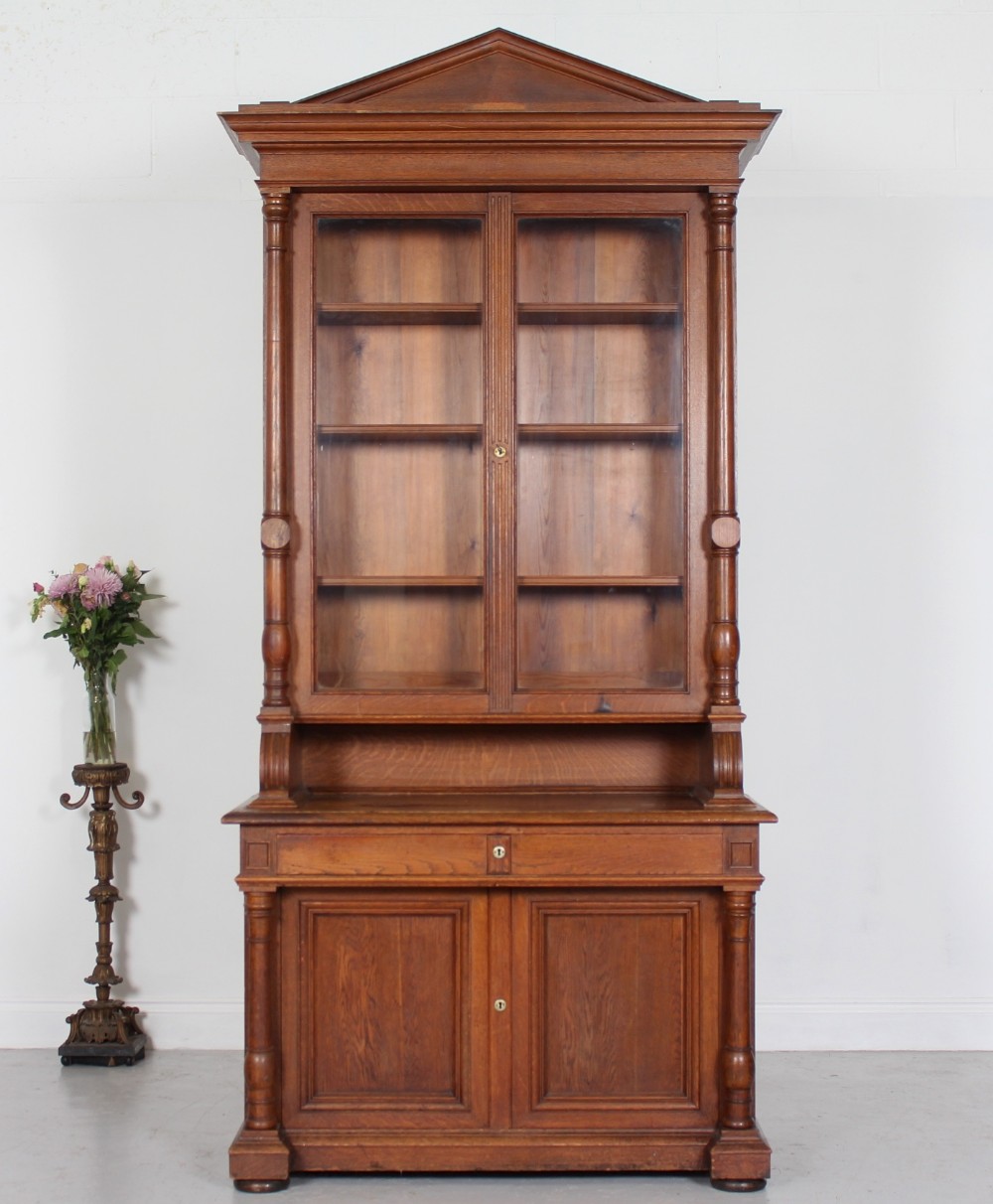 A tall nineteenth-century oak bookcase with a triangular pediment top, two glazed glass doors, a single middle drawer, and a lower double-door cupboard flanked by turned side columns.