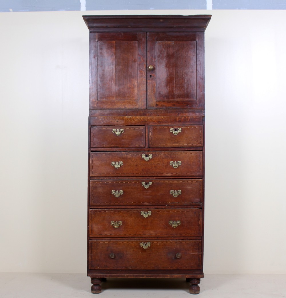 A tall 18th-century Georgian dark oak cupboard featuring panelled upper doors above six drawers with brass drop handles and stringing inlay, resting on turned feet.
