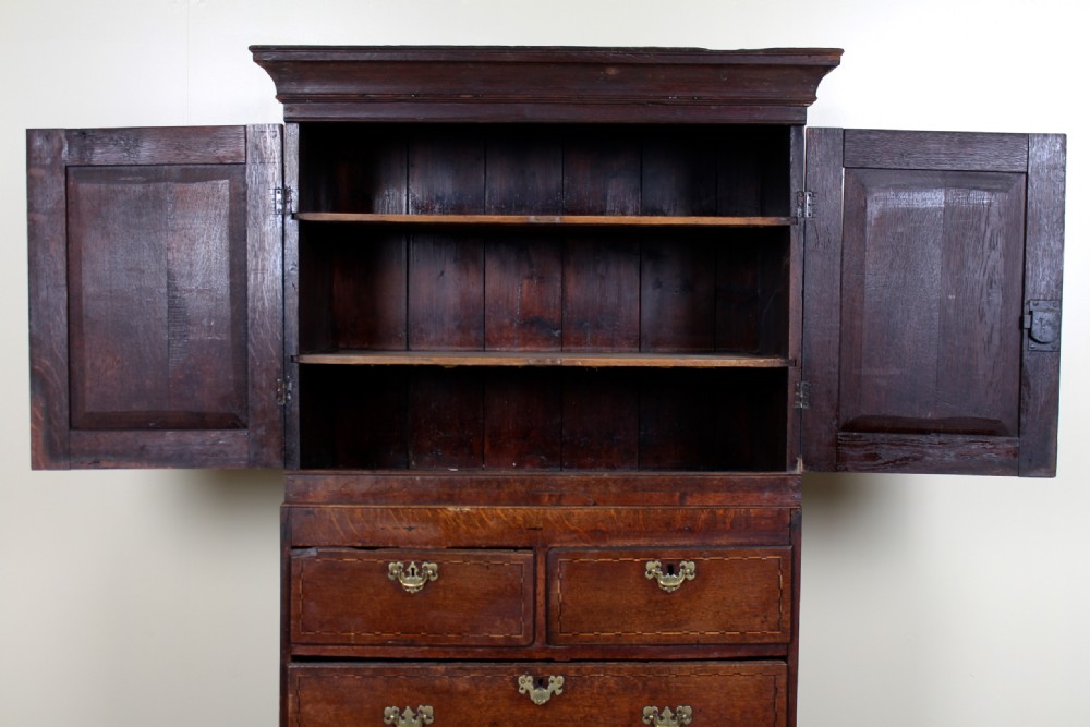 A tall 18th-century Georgian dark oak cupboard featuring panelled upper doors above six drawers with brass drop handles and stringing inlay, resting on turned feet. View V