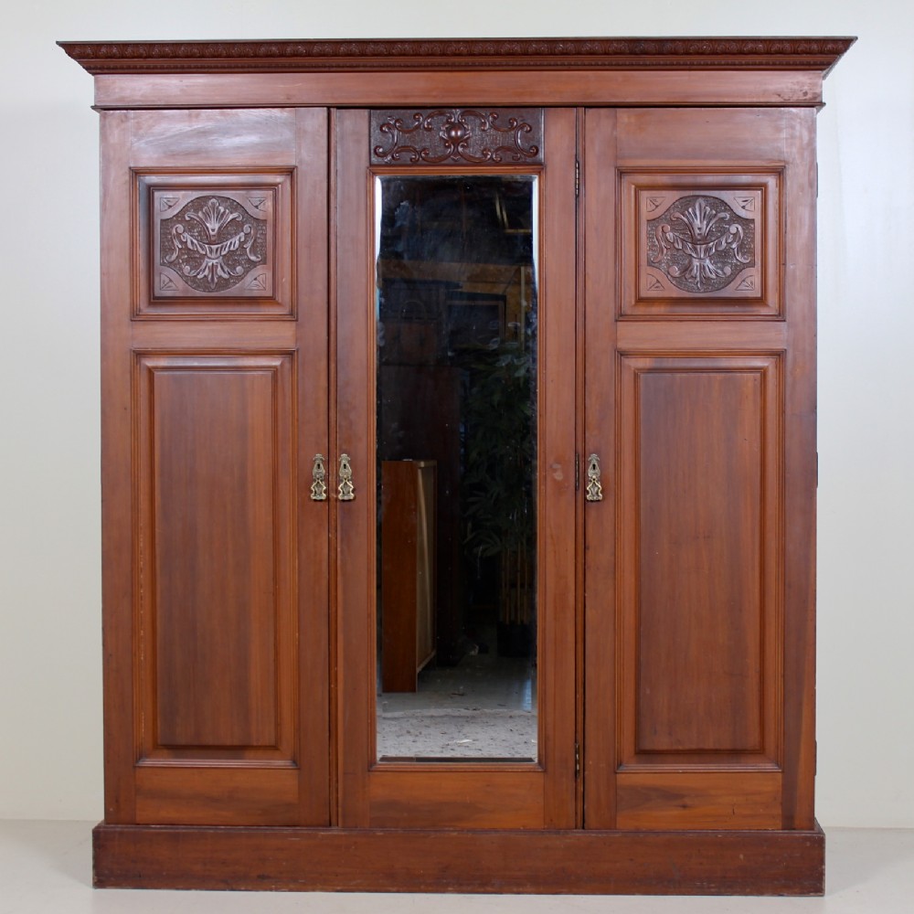 A Victorian triple-door mahogany wardrobe featuring a central mirrored door, carved foliate upper panels, and a solid plinth base.