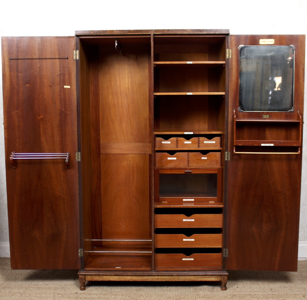 An open two-door Edwardian oak compactum wardrobe revealing a left hanging rail, right shelving, labelled drawers, a glass-fronted drawer, and an internal door mirror.