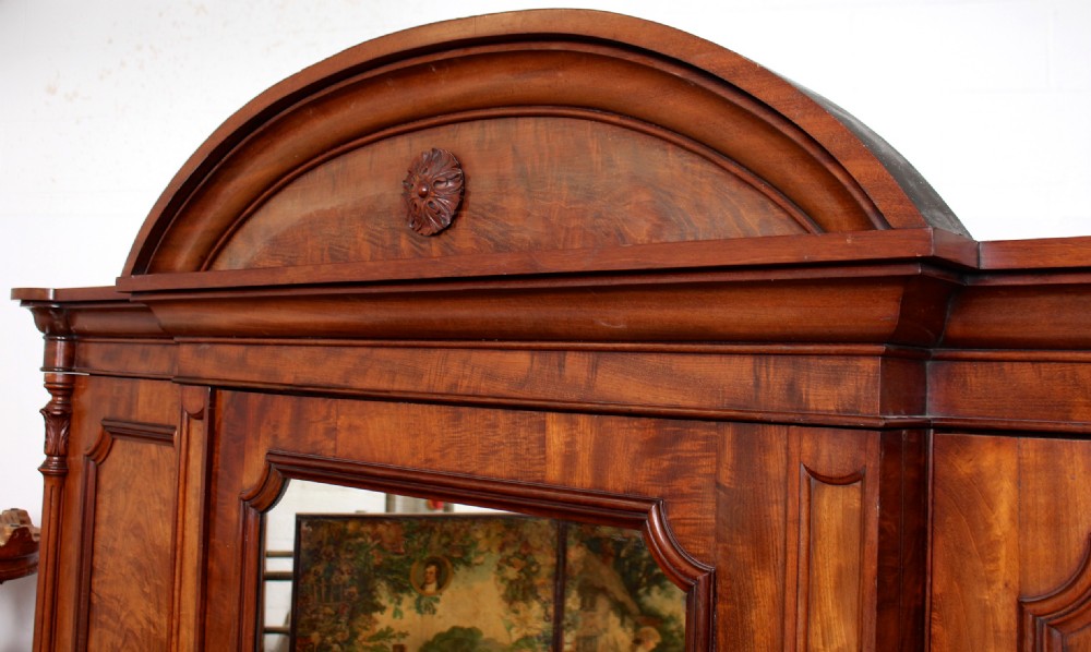 A 19th-century Victorian mahogany triple compactum wardrobe featuring a central mirrored door flanked by two flame veneer doors and topped with an arched pediment. View X
