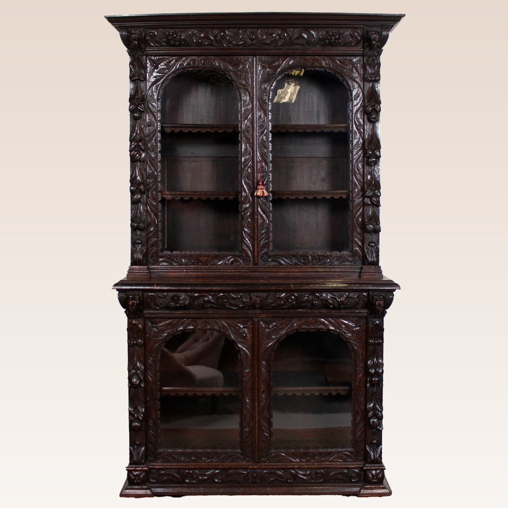 A two-tier dark brown ebonised oak Gothic Revival bookcase with carved foliate frames and arched glazed doors.