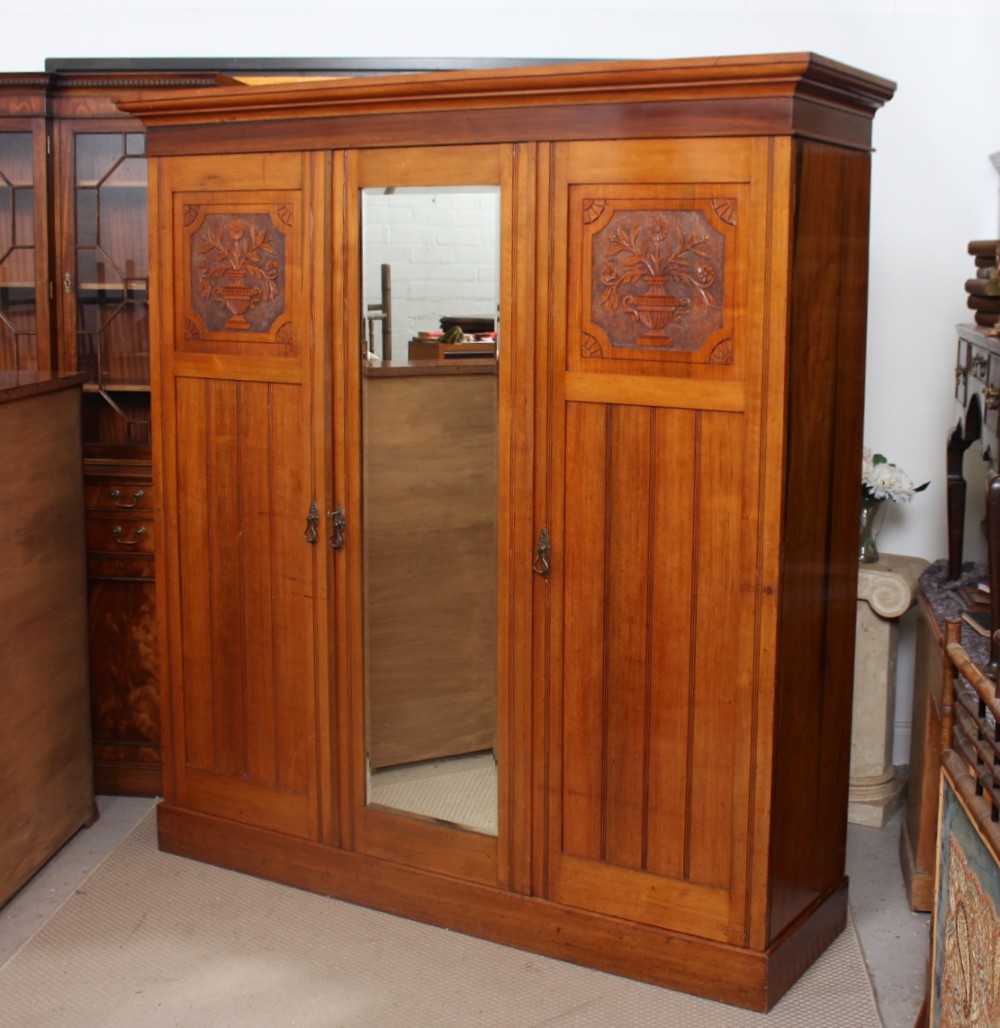 A large three-door walnut wardrobe featuring relief-carved floral urn panels on the outer doors and a tall central mirrored door.