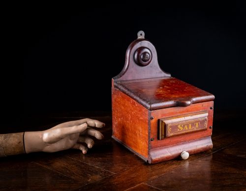 fig 1. A nineteenth-century reddish-brown painted wooden wall-hanging salt box featuring gilt lettering reading SALT, a sloping hinged lid, and a small bottom drawer.