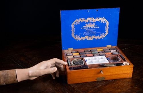 fig 2. A Victorian mahogany artist watercolour box featuring an open lid lined with blue gilt-stamped paper, fitted compartments containing colour cakes, glass dishes, and a lower drawer holding a ceramic palette.