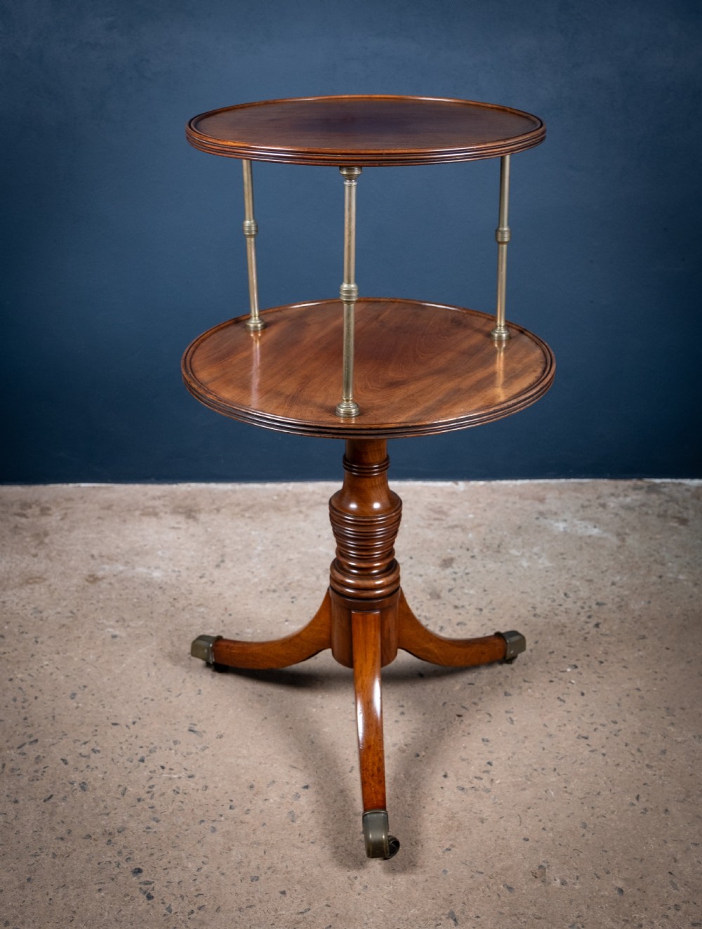 A two-tier Regency mahogany dumb waiter featuring a turned central column, brass uprights, and a tripartite base with castors. View II