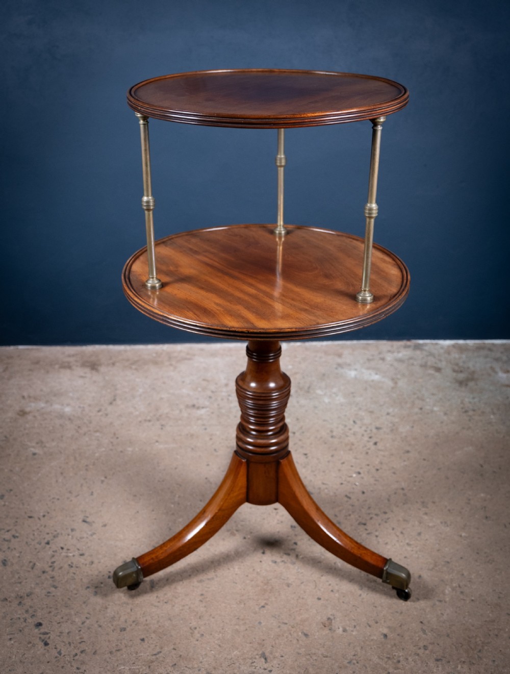 A two-tier Regency mahogany dumb waiter featuring a turned central column, brass uprights, and a tripartite base with castors.