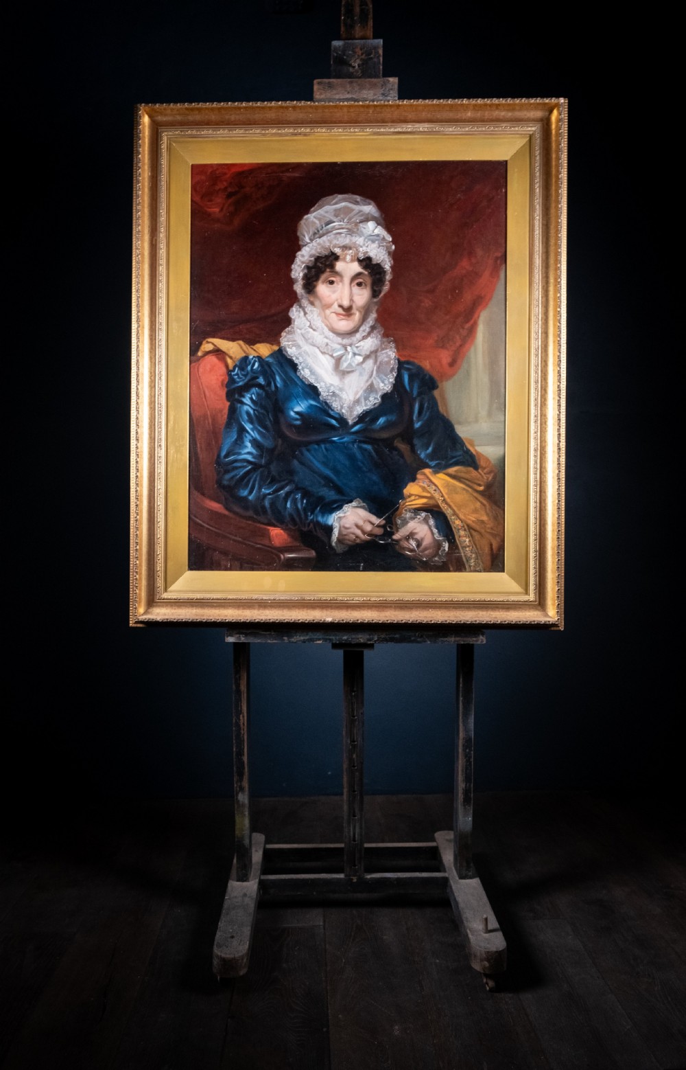 A large antique framed oil painting depicting a seated woman wearing a blue dress and white lace cap against a dark red backdrop. View IX