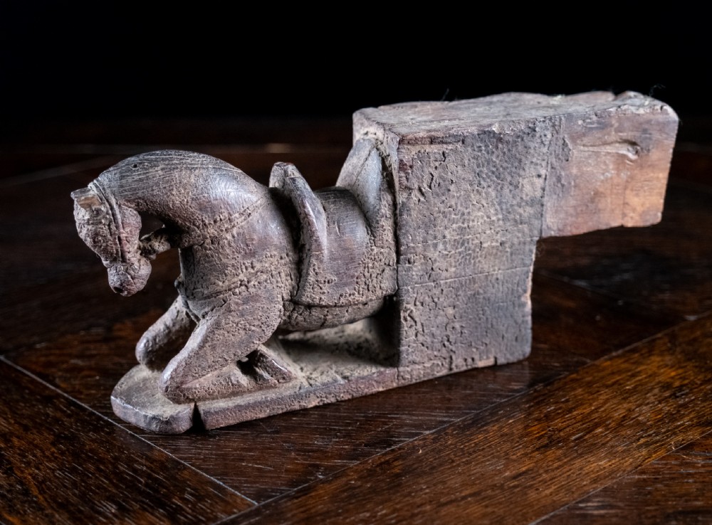 A close-up photograph of a weathered, dark brown carved wooden horse architectural element shown from a three-quarters front angle against a black background. View II