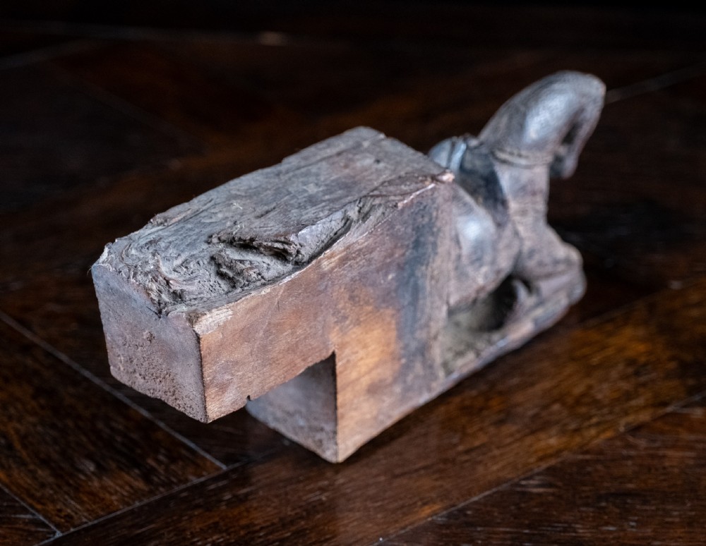 A close-up photograph of a weathered, dark brown carved wooden horse architectural element shown from a three-quarters front angle against a black background. View IV